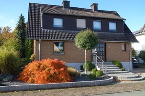 Une maison à deux étages, avec un toit en tuiles, une façade en briques et un petit jardin devant, agrémenté d’un arbre et de quelques arbustes.
