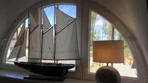 En modellsegelbåt står på en böjd fönsterbräda, bredvid en lampa, och utanför syns träd och himmel.