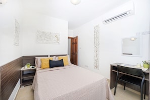 A bedroom with a bed, bedside table, desk, chair, and an air conditioning unit.