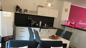 A modern living room with a dark grey sofa, a pink blanket, and a remote control on it. There is a dining area with a table and chairs, and a kitchen with white cabinets and a black countertop.