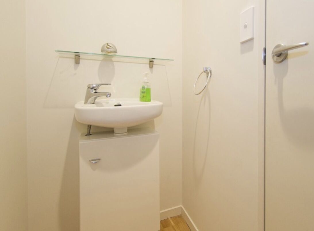 Een kleine badkamer met een witte wastafel, een glazen plank erboven en een douchehoek aan de rechterkant.