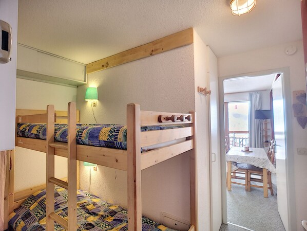 A bunk bed room with a wooden headboard, a green wall lamp, and a view of a dining area with a table and chairs.