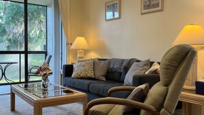 A living room with a ceiling fan, a sofa, a chair, two lamps, a coffee table, and a window with a view of trees. - Beautiful Florida Condo near beach in Bradenton (Bradenton)