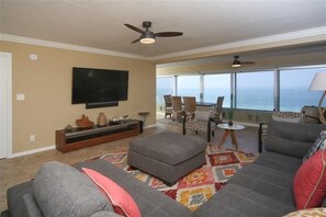 A living room with a large TV, a ceiling fan, a sofa, and a view of the ocean. - Horizons West #605 - Panoramic View on the #1 Beach (Sarasota)
