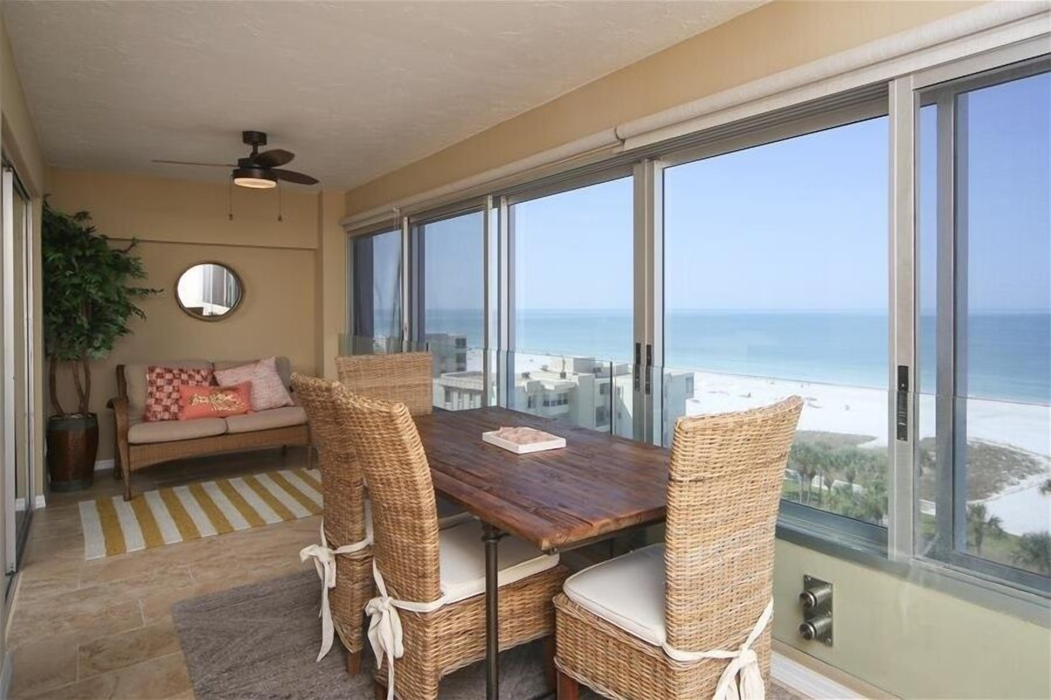 A dining area with a wooden table and wicker chairs, a ceiling fan, a sofa with cushions, and a large window overlooking a coastal view.