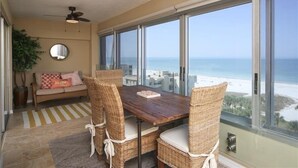 Un comedor con una mesa de madera y sillas de mimbre, un ventilador de techo, un sofá con cojines y un ventanal con vistas a la costa.