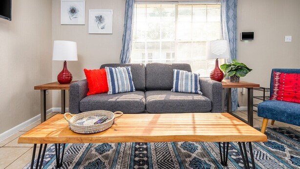 Uma sala de estar com um sofá cinza, almofadas listradas em vermelho e azul, uma mesa de centro de madeira, um vaso vermelho e uma cadeira azul.