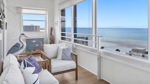 Un salon moderne doté d’une grande fenêtre donnant sur une plage, d’une chaise en bois et d’une commode aux couleurs vives, décorée de motifs de poissons.