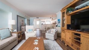 A living room with a wooden coffee table, white armchairs, a sofa, and a TV stand.
