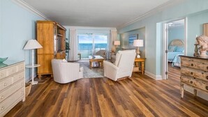 A spacious room with a wooden floor, a white armchair, a small table, and a view of the outdoors through a large window.