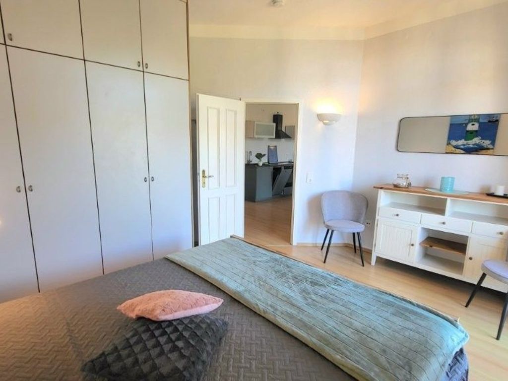 A bedroom with a bed, a desk, and a chair. There is a door leading to a kitchen area.