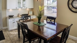 A kitchen with white cabinets, a dining table with chairs, a vase with plants, and a clock on the wall. - Horizons West Villa A-11 - on Siesta Key's #1 Beach (Sarasota)
