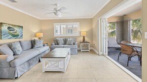 A living room with a sofa, coffee table, and a ceiling fan. There is a window with shutters and a view of the outdoors.