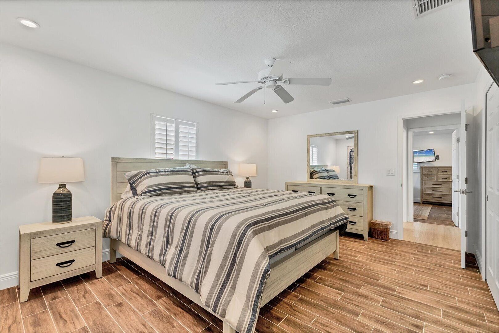 A bedroom with a bed, two bedside tables, a ceiling fan, a window, and a door leading to another room.