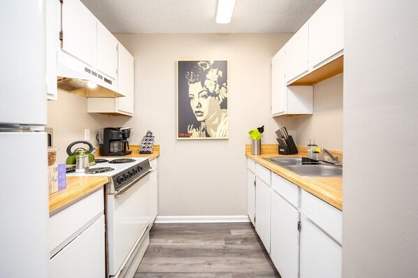 Cocina compacta con armarios blancos, encimera de madera, estufa, lavabo y un cartel en la pared.