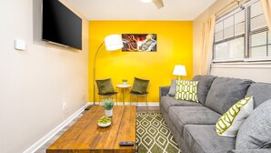 A living room with a yellow wall, a gray sofa, a wooden coffee table, two chairs, a TV, and a ceiling fan.