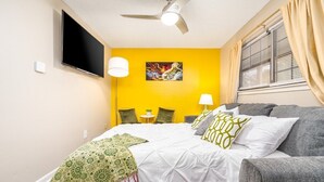 A bedroom with a bed, a grey sofa, a ceiling fan, a TV mounted on the wall, and a window with curtains.