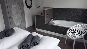 A bedroom with a bed, rolled towels, a grey wardrobe, a bathroom with a bathtub, and a white chair.