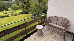 A balcony with a wicker chair and a small round table, overlooking a well-maintained garden with a wooden fence.