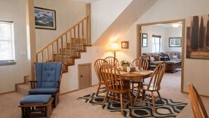 A dining area with a wooden table and chairs, a blue upholstered chair, a staircase, and a living room in the background. - Mountain Hideaway (Ruidoso)