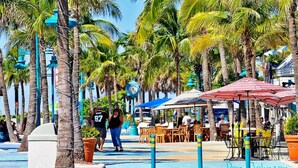 Calle bordeada de palmeras y zonas de estar al aire libre, probablemente un espacio público o cafetería.