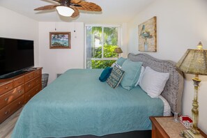 A bedroom with a bed, a television, a ceiling fan, a window with a view of greenery, and a painting on the wall.