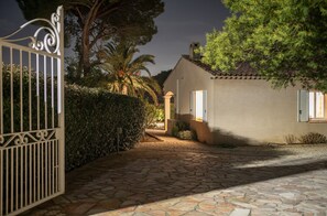 Un sendero de piedra que lleva a una casa con entrada protegida por una reja, rodeada de vegetación y una palmera.