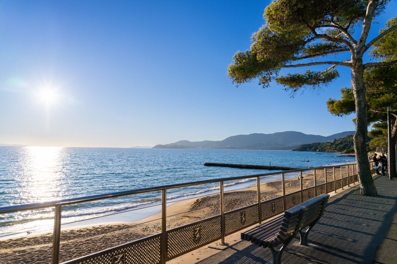 A coastal walkway with a bench, a railing, and a clear view of the sea and distant mountains.