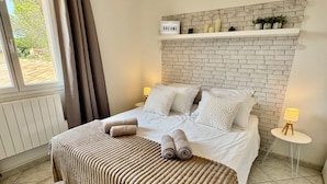 A bedroom with a bed, a headboard, a window with curtains, a radiator, a wall-mounted shelf with decorative items, and a bedside table with a lamp.