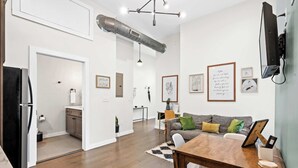 A modern living room with a wooden dining table, grey sofa with green and yellow cushions, and a kitchen area in the background. - 4 Apartments, Downtown, Walk to Cincy Stadiums! (Covington)