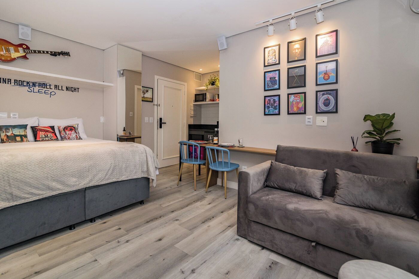 Een moderne slaapkamer met een bed, een grijze bank, een bureau en een stoel. Er hangen afbeeldingen in lijsten aan de muur en een gitaar.
