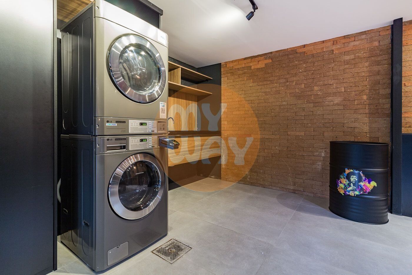 A laundry area with stacked washing machines, a brick wall, and a black barrel with a colorful design.