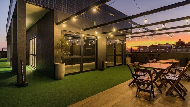 An outdoor patio with tables and chairs, a green artificial turf area, and a building with large windows and string lights. - Century Park by My Way (Porto Alegre)