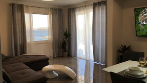 A modern living room with a sofa, a white ottoman, a dining table with chairs, and a view of the sea through large windows. - Holiday Hvar LUX 2.  (Vrbanj)