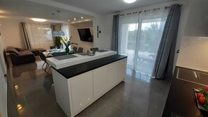 A modern kitchen with a central island, a dining area with a sofa and table, and a large window with curtains. - Holiday Hvar LUX 2.  (Vrbanj)