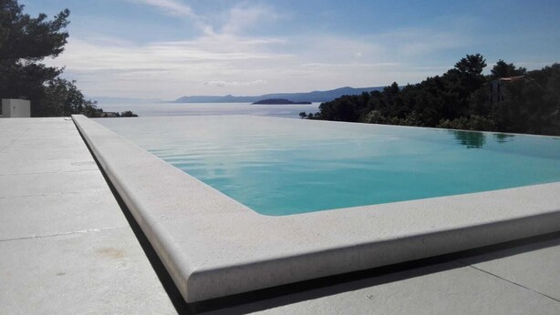 A rectangular infinity pool with a clear blue edge, surrounded by a light-colored deck, set against a backdrop of trees and distant mountains under a partly cloudy sky. - Holiday Hvar LUX 2.  (Vrbanj)
