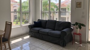 Un salon avec un canapé gris, une petite table et un fauteuil. De grandes fenêtres offrent une vue sur un quartier résidentiel.
