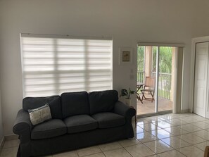 A living room with a dark grey sofa, a potted plant, and a sliding glass door leading to an outdoor patio area. - Condo in PGA Village Mets Spring Training (Port St. Lucie)