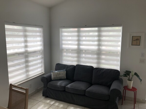 A living room with a grey sofa, two windows with blinds, a small red stool with a plant, and a framed picture on the wall. - Condo in PGA Village Mets Spring Training (Port St. Lucie)