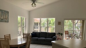 Un vaste espace de vie comprenant une table à manger, des chaises, un canapé et un ventilateur de plafond. De grandes fenêtres laissent entrer la lumière naturelle et offrent une vue sur l’extérieur.