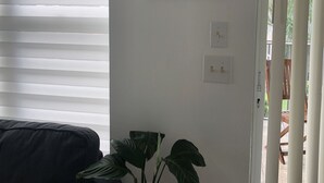 Une pièce avec un canapé aux couleurs sombres, une plante en pot posée sur un petit tabouret, une œuvre d’art encadrée accrochée au mur et une porte coulissante donnant sur un espace extérieur.