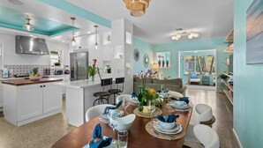 Una cocina moderna con armarios blancos, una isla central y un comedor con una mesa de madera dispuesta para seis personas.