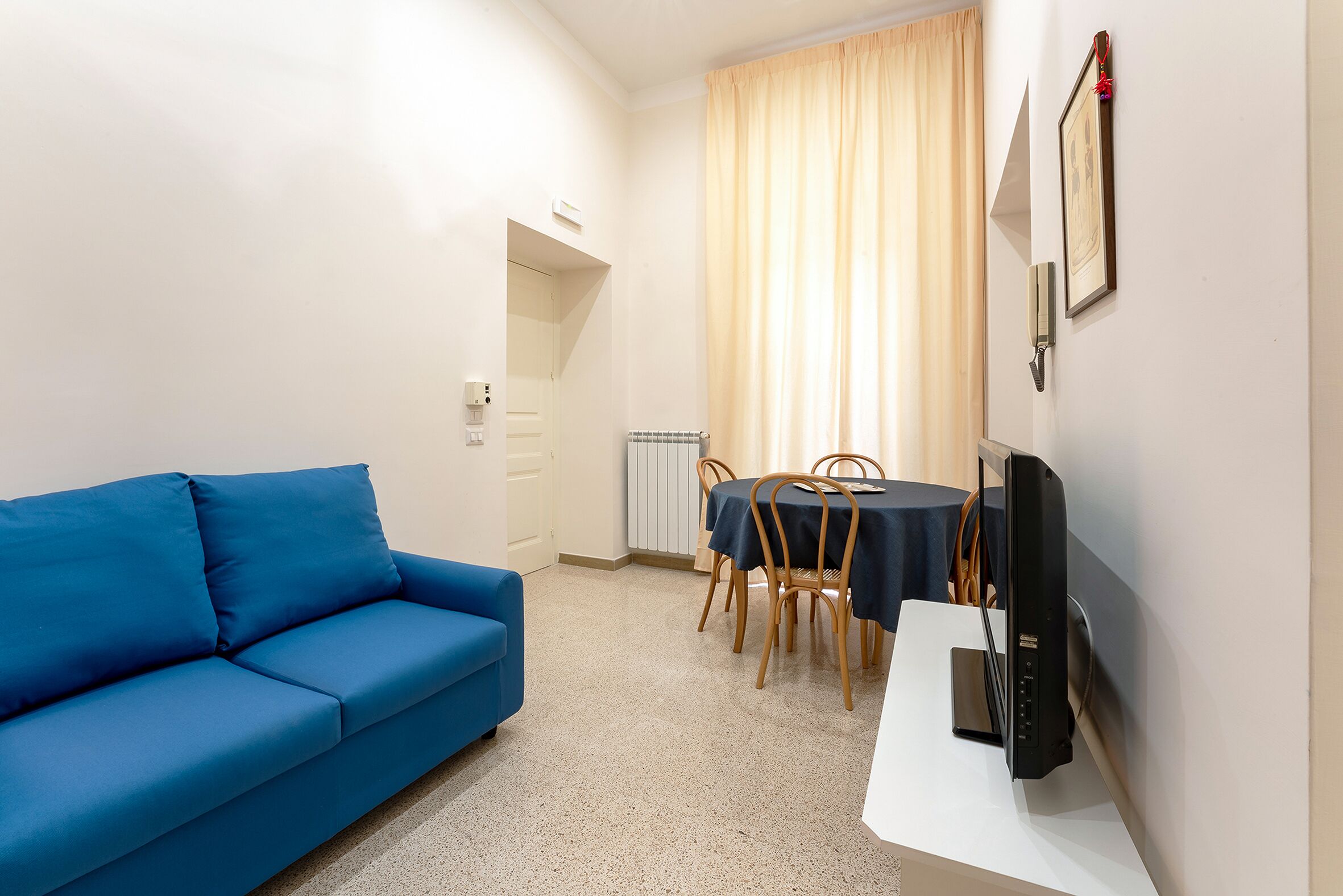 Ein Zimmer mit einem blauen Sofa, einem Esstisch mit Stühlen und einem Flachbildfernseher auf einem Lowboard.