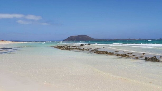 Una playa con aguas turquesas cristalinas, una orilla arenosa y una montaña distante.