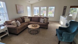 A living room with a brown sectional sofa, a round coffee table, a blue armchair, and a small white side table with a lamp. - Pet Friendly Home with Private Tiki Bar - 3-minute drive to the beach! (Kill Devil Hills)