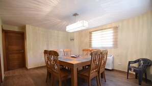 A dining area with a wooden table and chairs, a ceiling light fixture, a window with blinds, and a door.