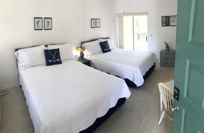 Habitación de hotel con dos camas, una mesita de noche con lámpara, una pequeña mesa con una silla y una puerta con cerradura electrónica.