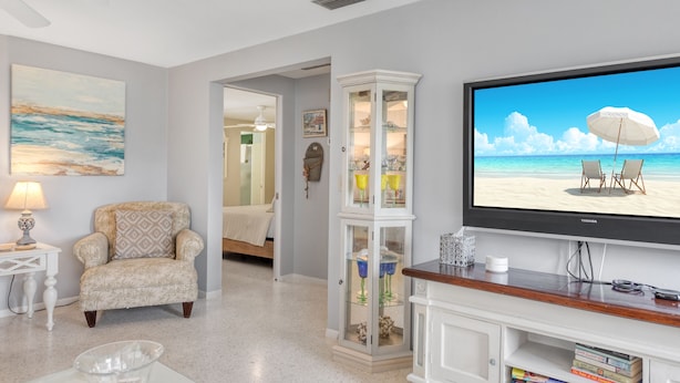 A living room with a television displaying a beach scene, a white armchair, a glass cabinet, and a small table with a lamp.