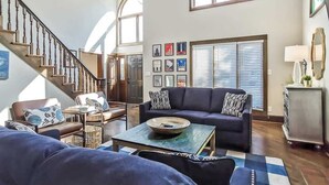 Un salon spacieux avec un escalier, un ensemble de canapés, une table basse et une grande fenêtre équipée de stores.