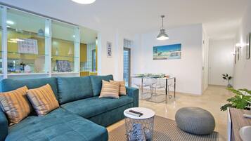 A modern living room with a sectional sofa, a dining area with a glass table, and a kitchen visible through a glass partition.